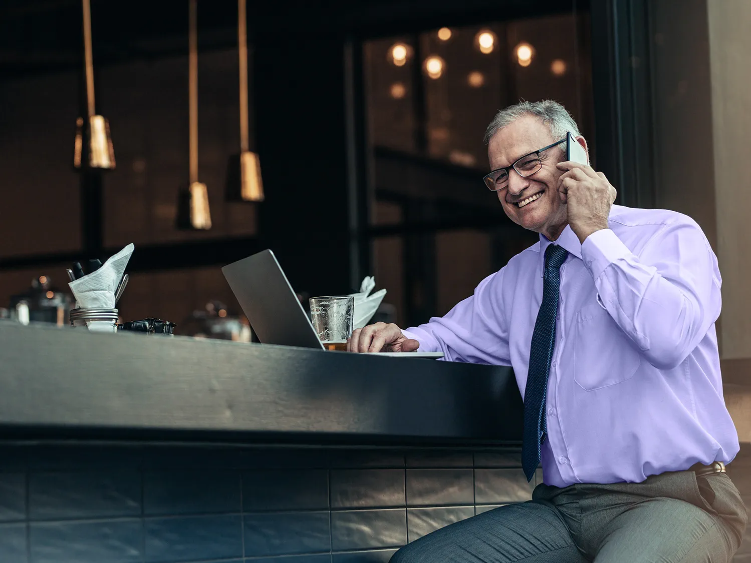 Smiling senior man in formal wear sitting at bar counter with laptop talking on cell phone wearing hearing aids