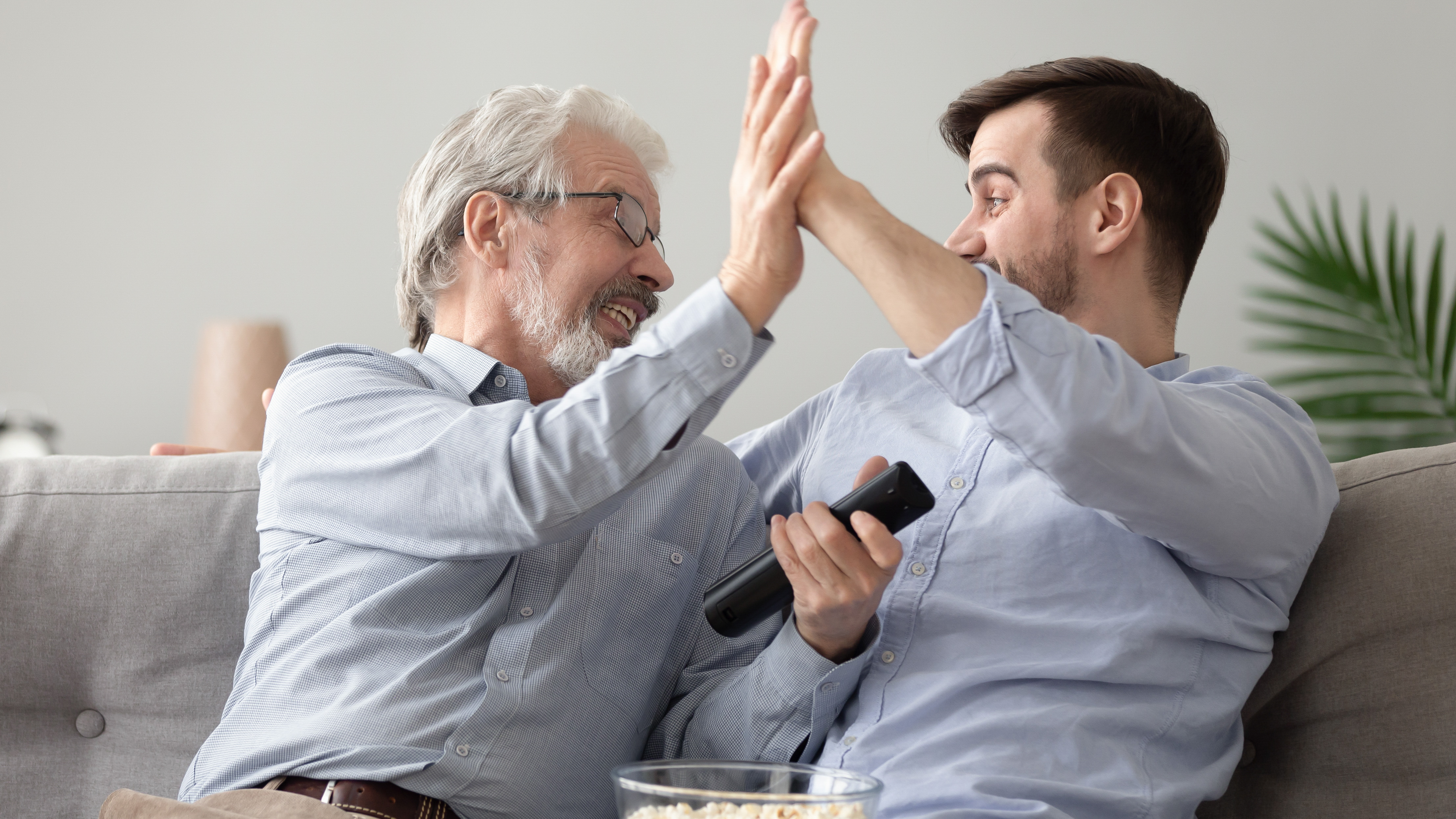 Happy elderly father and millennial son sit on couch give high five celebrating team win online game, excited young man and senior dad feel euphoric after watching TV football match together at home using hearing aids