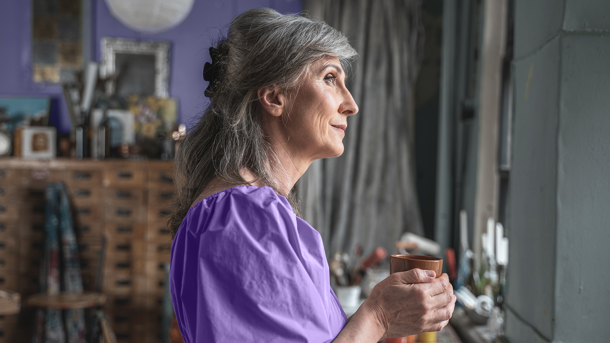 A woman holding a cup of tea stands in her kitchen, contemplating her hearing health
