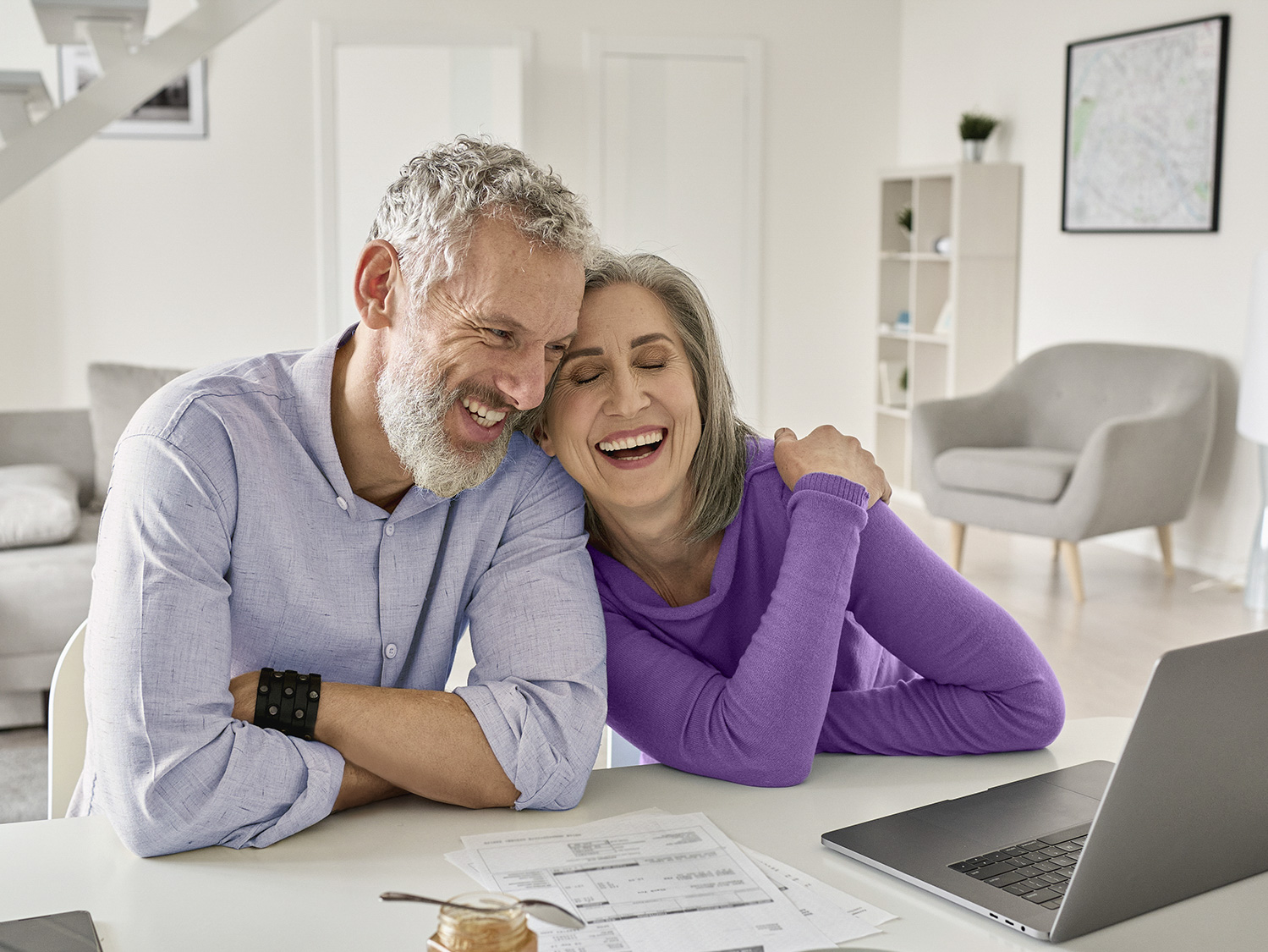 Happy mature couple sitting at home table with laptop wearing hearing aids