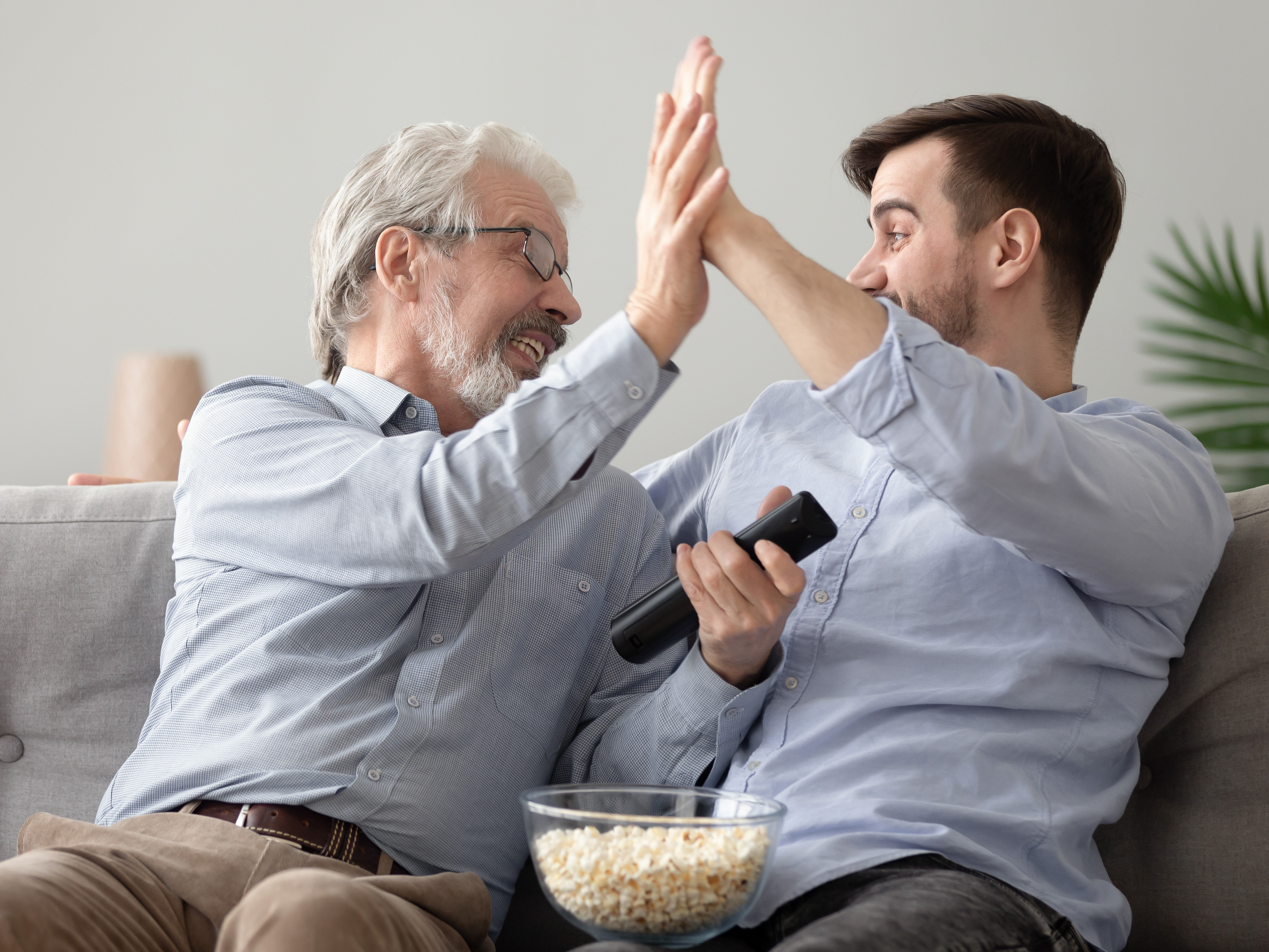 Happy elderly father and millennial son sit on couch give high five celebrating team win online game, excited young man and senior dad feel euphoric after watching TV football match together at home using hearing aids